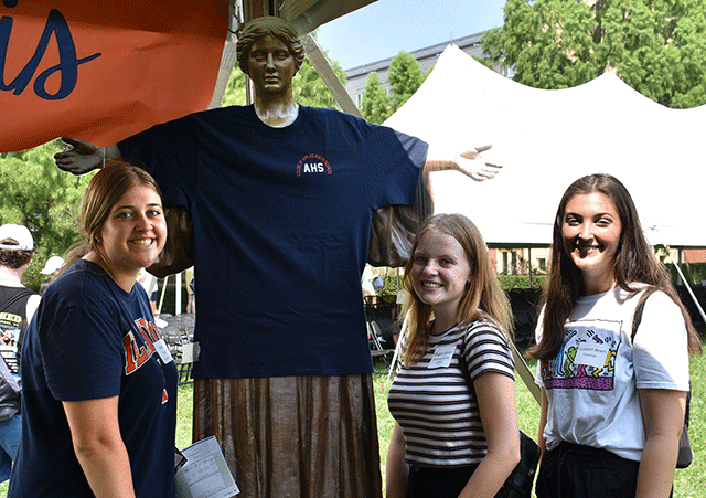 three young women standing with cardboard figure of Alma Mater