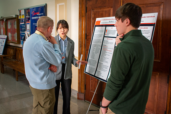 A man in a blue sweater listens to students give a research poster presentation.