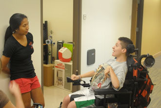 student in powered chair talking to a student standing against a wall